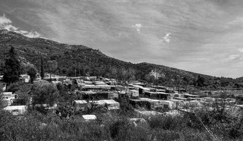 Black and  landscape image Samos. Grass, trees, a mountain and blue sky frame a large camp made up of huts.