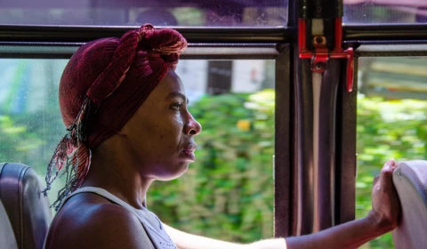 A photograph of a black woman with a red scarf tied over her head, sitting on a bus, looking forward with a serious expression.