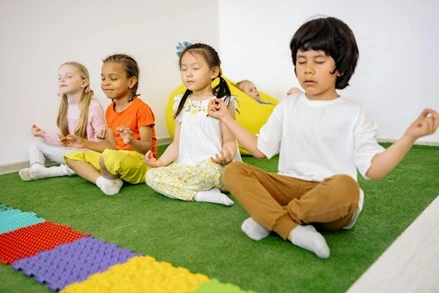 A diverse group of four children sit in a white room on artificial grass. Each of them sits with crossed legs and eyes closed in a lotus position.