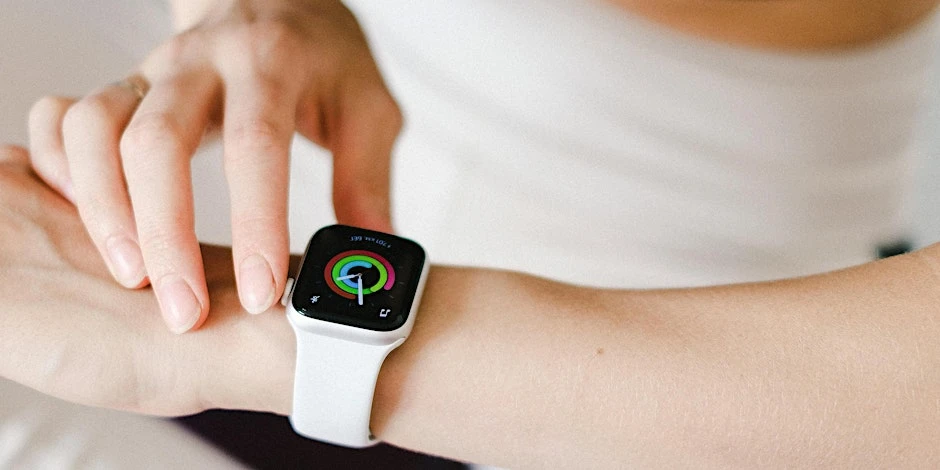 The arm of a slim, white woman wearing a smart watch, with the other hand reaching towards it.