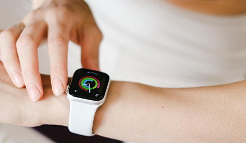 The arm of a slim, white woman wearing a smart watch, with the other hand reaching towards it.