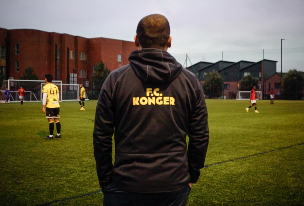 Photograph of someone watching a grassroots football match. They are wearing a Konger FC hoodie.