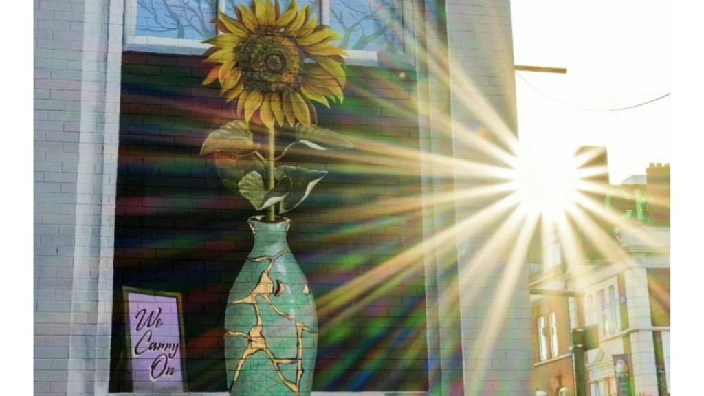 A mural of a sunflower in a kintsugi-style vase with the words “We Carry On,” lit by sunbeams, symbolizing hope and resilience.