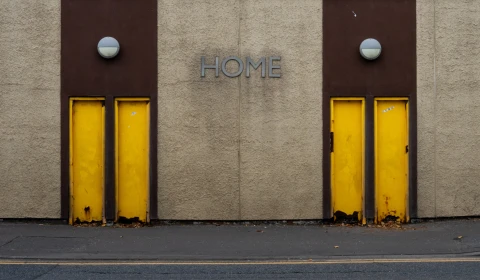 A picture of four yellow doors on a brown sandstone building. A sign situated in the middle, also on the wall of the building, says 'Home'