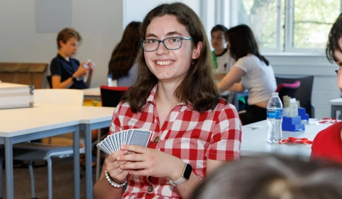 An image of a girl with shoulder length brown hair and blue glasses wearing a red checked shirt. She is smiling and holding cards in her hand.