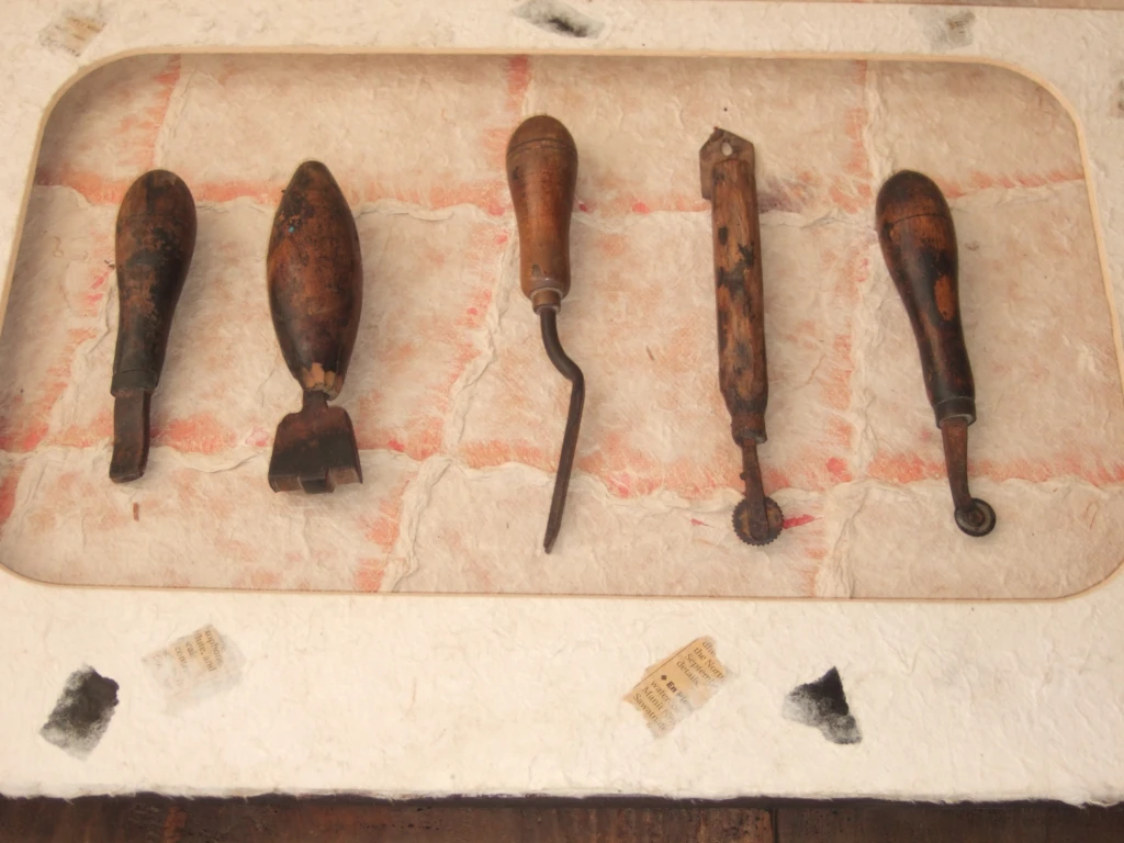 A display of five old, worn wooden-handled tools is laid out horizontally against a pale textured background with faint red grid-like lines. The tools are of different shapes and appear to be traditional hand tools, possibly used for leatherwork.