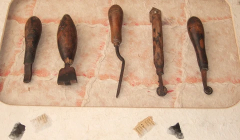 A display of five old, worn wooden-handled tools is laid out horizontally against a pale textured background with faint red grid-like lines. The tools are of different shapes and appear to be traditional hand tools, possibly used for leatherwork.